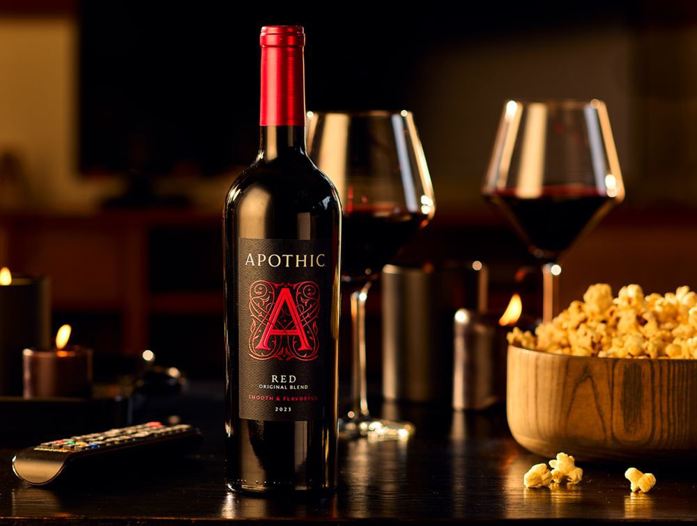 A warm, high-contrast lifestyle photograph of a movie night at home. In the center, a bottle of 2023 Apothic Red Original Blend sits on a dark wooden table next to two filled wine glasses. To the right, a large wooden bowl is filled with popcorn. In the foreground, a television remote rests on the table, while glowing candles in the background create a cozy, ambient atmosphere.