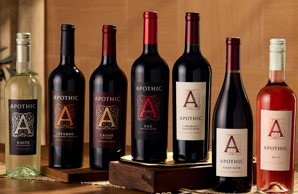 A wide, cinematic shot of ten diverse bottles of Apothic wine arranged in a line on a rustic wooden table. The collection includes Apothic Dark, White, Inferno, Crush, Red, Cabernet Sauvignon, Pinot Noir, Ros&eacute;, Merlot, and Chardonnay. A gold corkscrew sits in the foreground, and the background features warm, soft-focus lighting that highlights the varied colors of the wine from deep reds to pale golds.