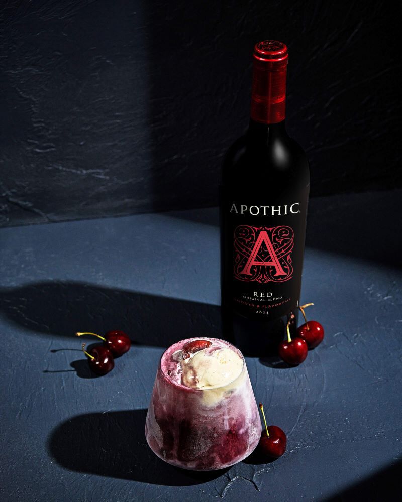 A moody lifestyle shot of a Red Float served in a stemless glass on a dark, textured surface. The glass is filled with a bubbly, deep-red wine and club soda mixture, topped with a large scoop of melting vanilla ice cream and a fresh cherry. To the right, a bottle of 2023 Apothic Red Original Blend stands against a dark wall. Three additional fresh cherries are scattered on the surface near the glass, emphasizing a high-contrast, atmospheric aesthetic.