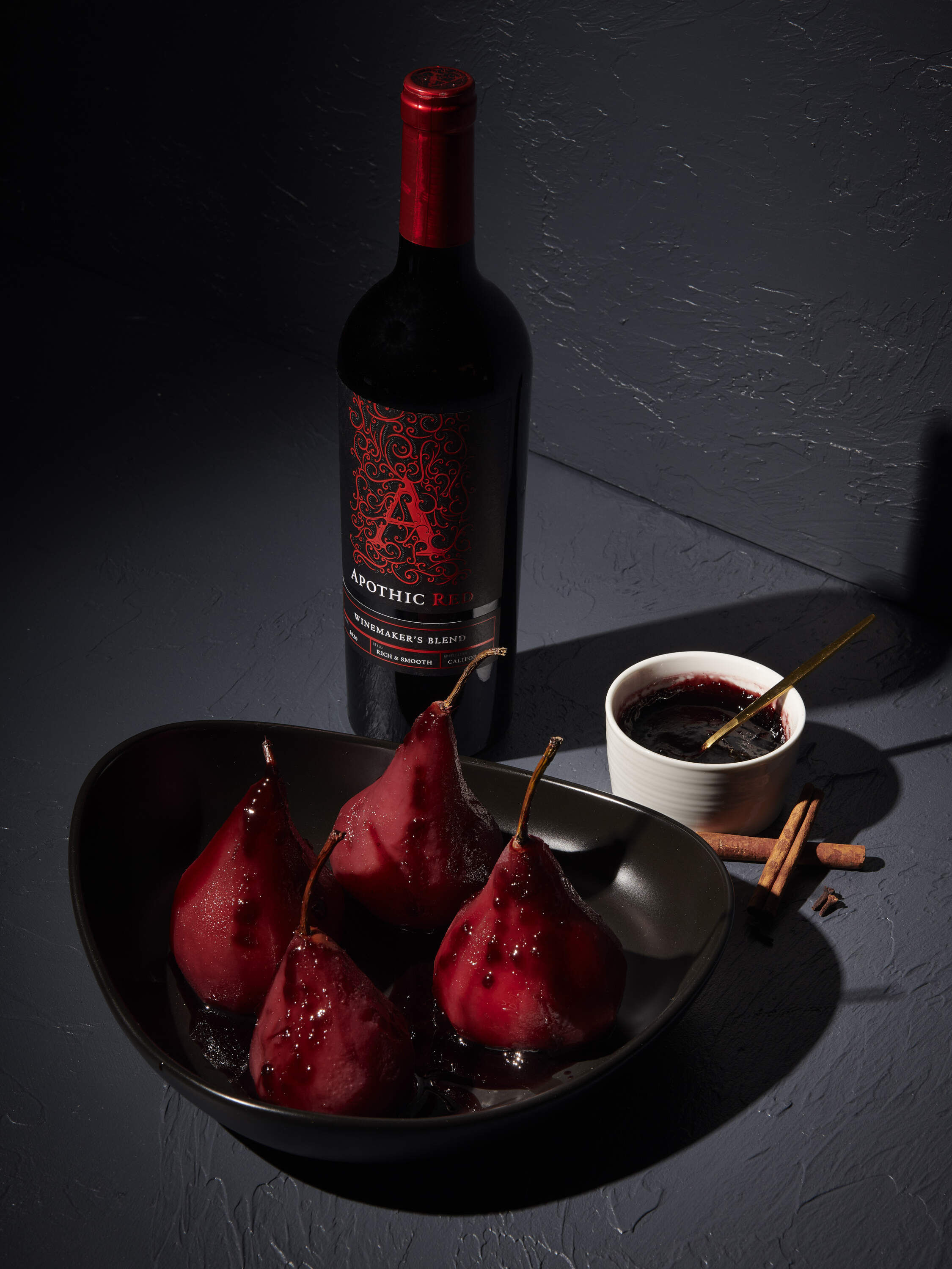 A sophisticated, dark-toned lifestyle photograph of two whole pears poached in red wine, served in a shallow dark bowl. The pears are a deep crimson color and are garnished with a cinnamon stick and a star anise pod. In the background, a bottle of Apothic Red Winemaker's Blend and a filled wine glass sit on a textured dark gray surface, creating a moody and elegant dessert presentation.