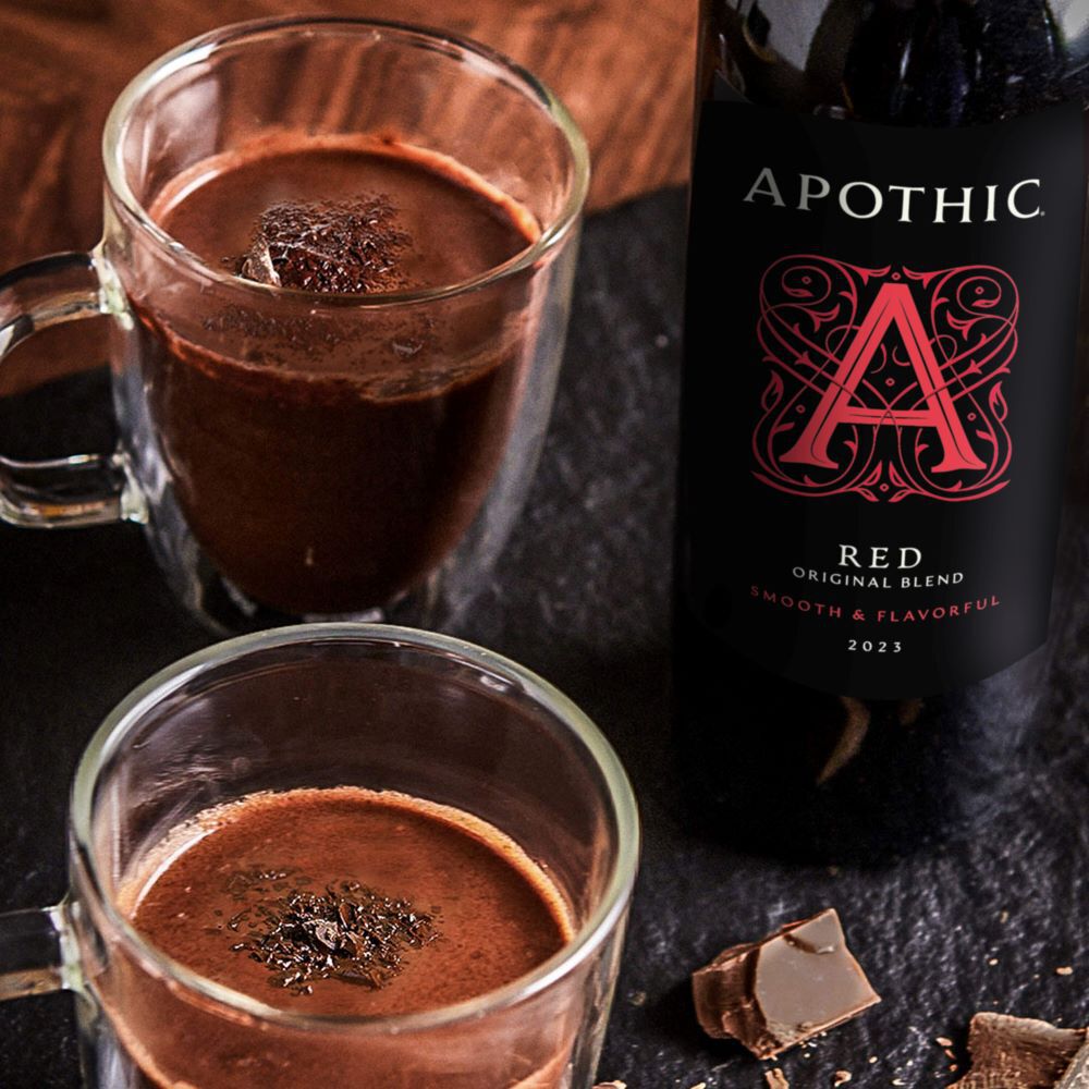 A warm, indulgent red wine hot cocoa served in a clear glass mug on a dark, textured surface. The drink is topped with a large toasted marshmallow and a square of dark chocolate. To the left, a bottle of Apothic Red Original Blend stands next to a small wooden bowl of dark chocolate chunks. The lighting is moody and focused, highlighting the steam and the rich texture of the chocolate and wine blend.