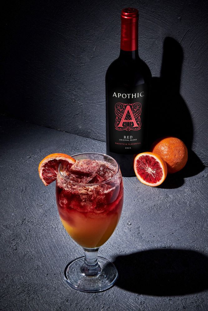 A bright, refreshing Red Twist cocktail served in a tall fluted glass on a dark, reflective surface. The ruby-red drink shows active carbonation bubbles and is garnished with an orange slice. To the left, a bottle of Apothic Red Original Blend stands next to a small wooden bowl containing fresh orange segments. The lighting is crisp and clear, emphasizing the effervescence of the club soda and the vibrant citrus accents.