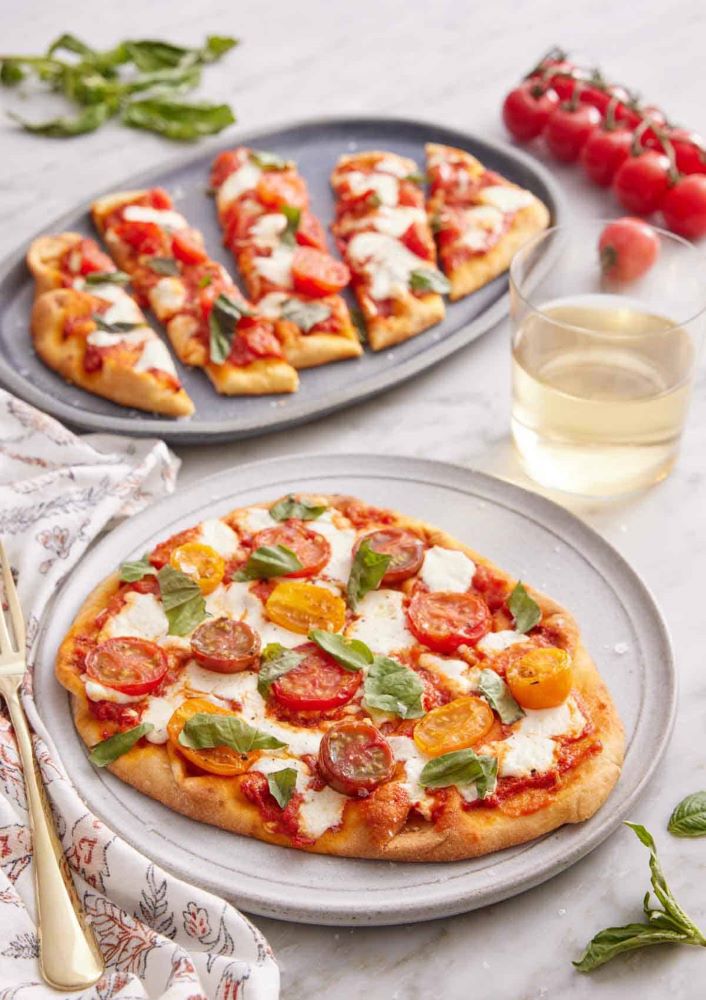 A bright, overhead lifestyle shot of a rectangular flatbread pizza topped with melted cheese, blackberries, and fresh herbs, sliced into pieces on a white marble surface. A bottle of 2022 Apothic Red Original Blend and two glasses of red wine are positioned at the top of the frame. To the left, a small wooden bowl contains fresh blackberries and mint leaves, emphasizing the unique fruit pairing.