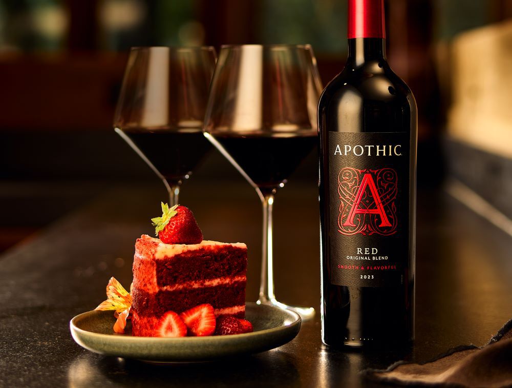 A close-up lifestyle photograph of a decadent date night pairing. A slice of rich red velvet cake topped with fresh strawberries sits on a small plate next to a bottle of 2023 Apothic Red Original Blend. Two filled wine glasses stand in the softly blurred background on a dark tabletop. The warm, amber lighting emphasizes the deep colors of the wine and the indulgent texture of the dessert.
