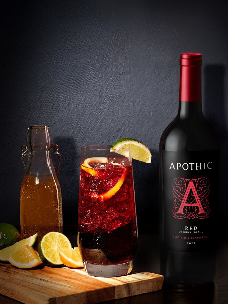 A refreshing red wine highball served in a tall, fluted glass on a dark, reflective surface. The drink is a deep ruby color with visible carbonation and is garnished with a fresh lemon wheel. To the left, a bottle of Apothic Red Original Blend stands next to a small wooden bowl containing lemon and lime wedges. The lighting is bright and crisp, highlighting the bubbles and the condensation on the glassware.