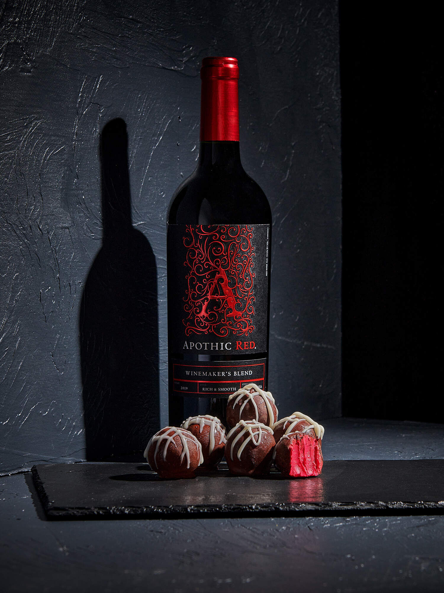 A sophisticated, dark-toned lifestyle photograph featuring six round chocolate truffles on a dark slate board. One truffle is bitten open to reveal a vibrant, deep pink red wine ganache center. Each truffle is drizzled with white chocolate. In the background, a bottle of Apothic Red Winemaker's Blend stands against a textured dark gray wall, casting a long shadow to create a moody and luxurious dessert scene.