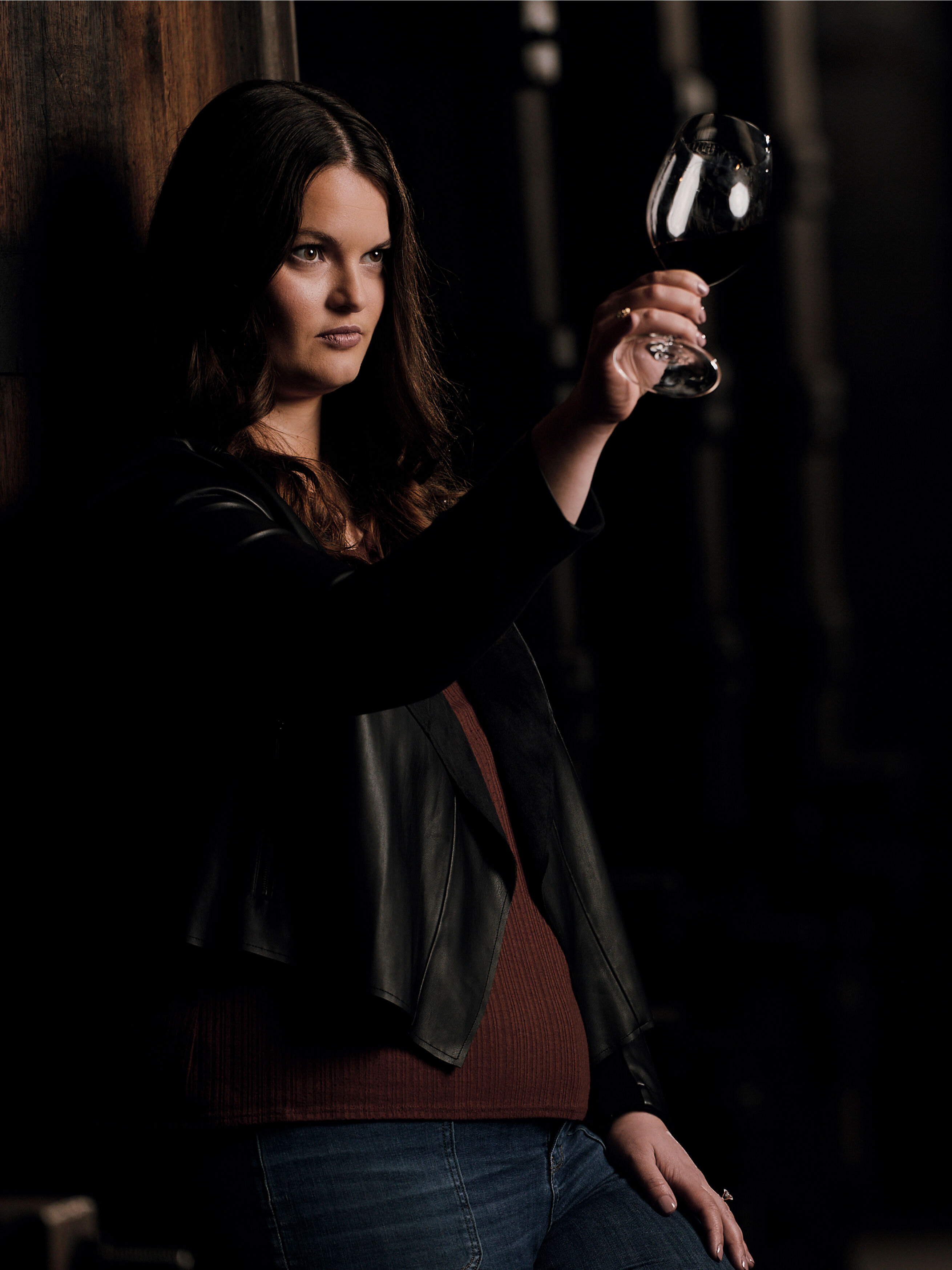 A moody, low-light portrait of Apothic Winemaker Ash Ricchio in a cellar setting. She is wearing a dark leather jacket over a maroon top and jeans, holding up a glass of red wine to the light to inspect its color. The background is deep and shadowy with vertical architectural elements, creating a sophisticated and focused atmosphere that highlights the artistry of winemaking.