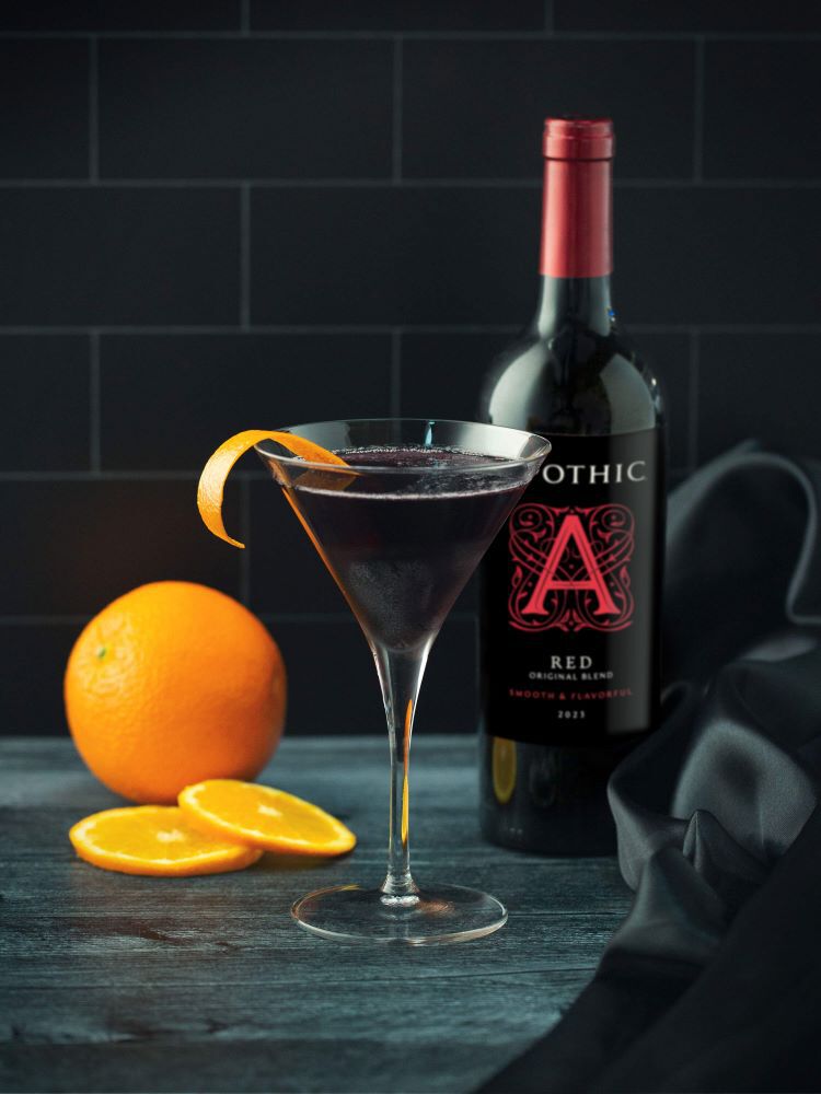 A vibrant Red Cosmo cocktail served in a chilled martini glass on a dark, reflective surface. The bright crimson drink is garnished with a thin, circular orange slice. In the background, a bottle of Apothic Red Original Blend stands next to a clear glass cocktail shaker and a small bowl of fresh orange segments. The lighting is crisp, highlighting the condensation on the glass and the deep color of the wine-based cocktail.
