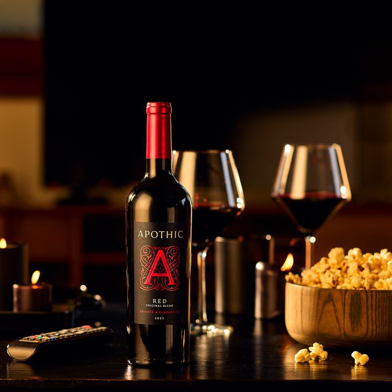 A warm, high-contrast lifestyle photograph of a movie night at home. In the center, a bottle of 2023 Apothic Red Original Blend sits on a dark wooden table next to two filled wine glasses. To the right, a large wooden bowl is filled with popcorn. In the foreground, a television remote rests on the table, while glowing candles in the background create a cozy, ambient atmosphere.