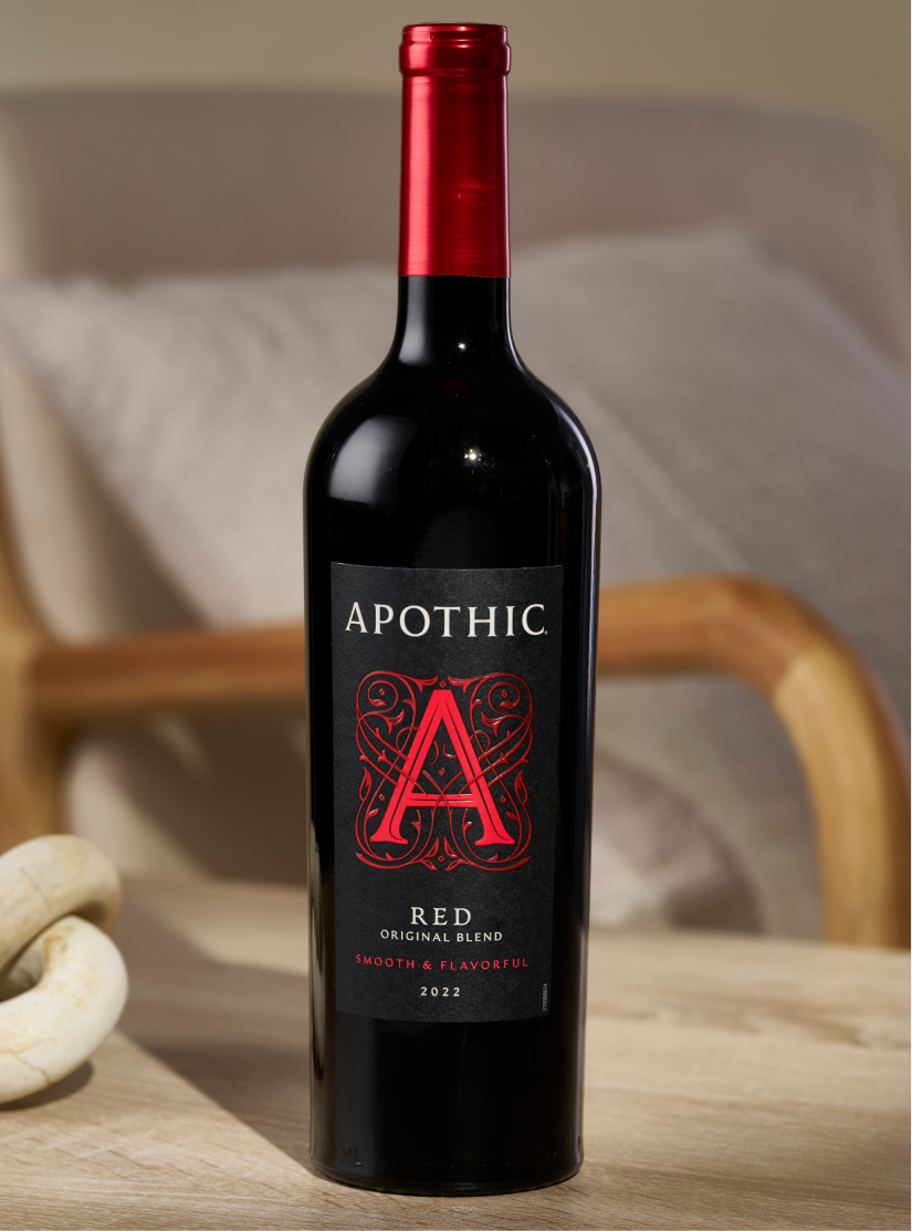A centered, close-up shot of a bottle of 2022 Apothic Red Original Blend standing on a light wooden surface. In the softly blurred background, a cream-colored armchair and a decorative wooden knot sit in a brightly lit room. The focus is sharp on the black label featuring the signature red 'A' logo, highlighting the 'Smooth and Flavorful' description.